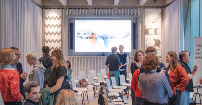 Auf dem Foto sind verschiedene Menschen in Gespräche vertieft zu sehen, am Ende des Raumes steht auf einer Leinwand: Her mit der Öffentlichkeitspolitik!
