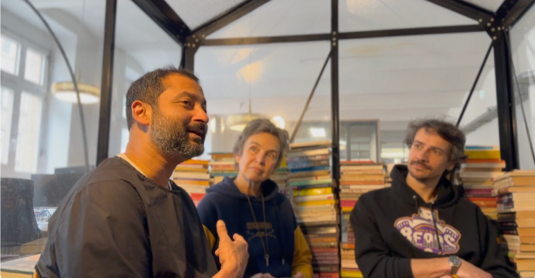 Aaron, Joana und Lukas sitzen in einem Glashaus mit vielen Büchern an der Wand.