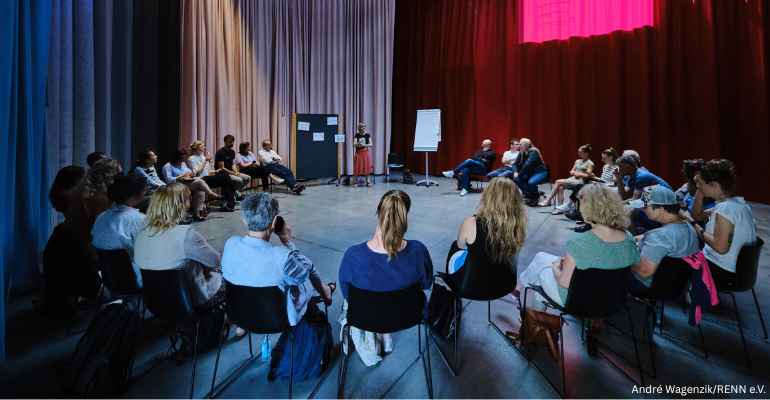 Ungefähr 20 Menschen sitzen in einem Stuhlkreis. Zwischen einer Pinnwand und einem Flipchart in der Mitte des Bildes steht eine Person und spricht.